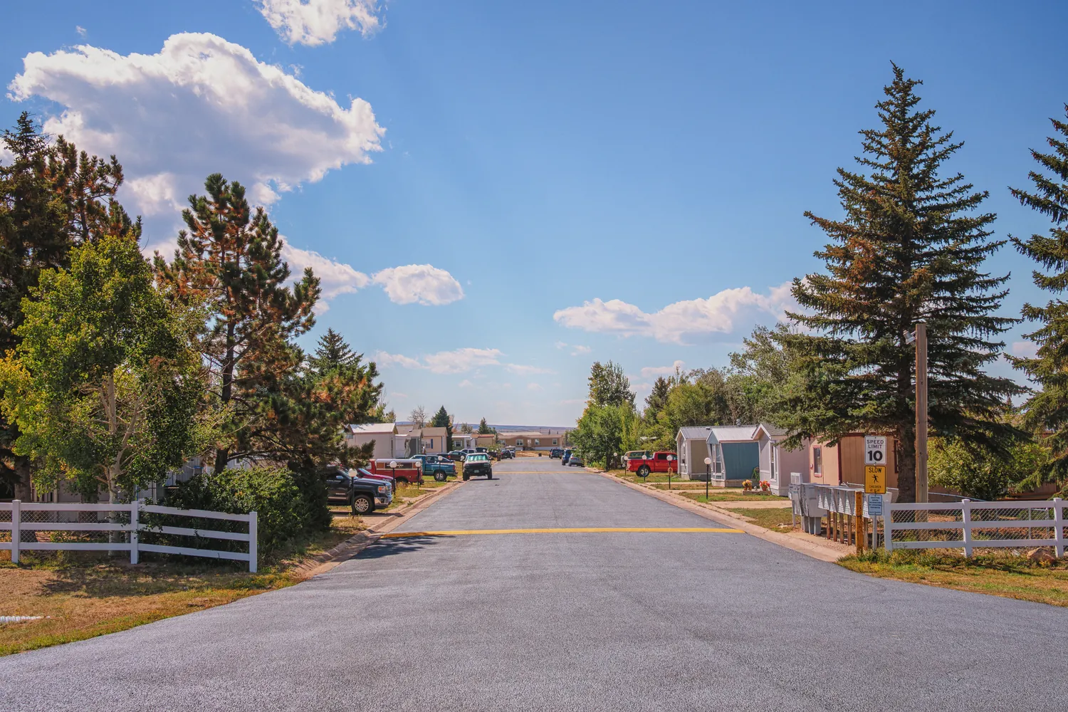 Paved entry road into a fenced, tree-lined mobile home community